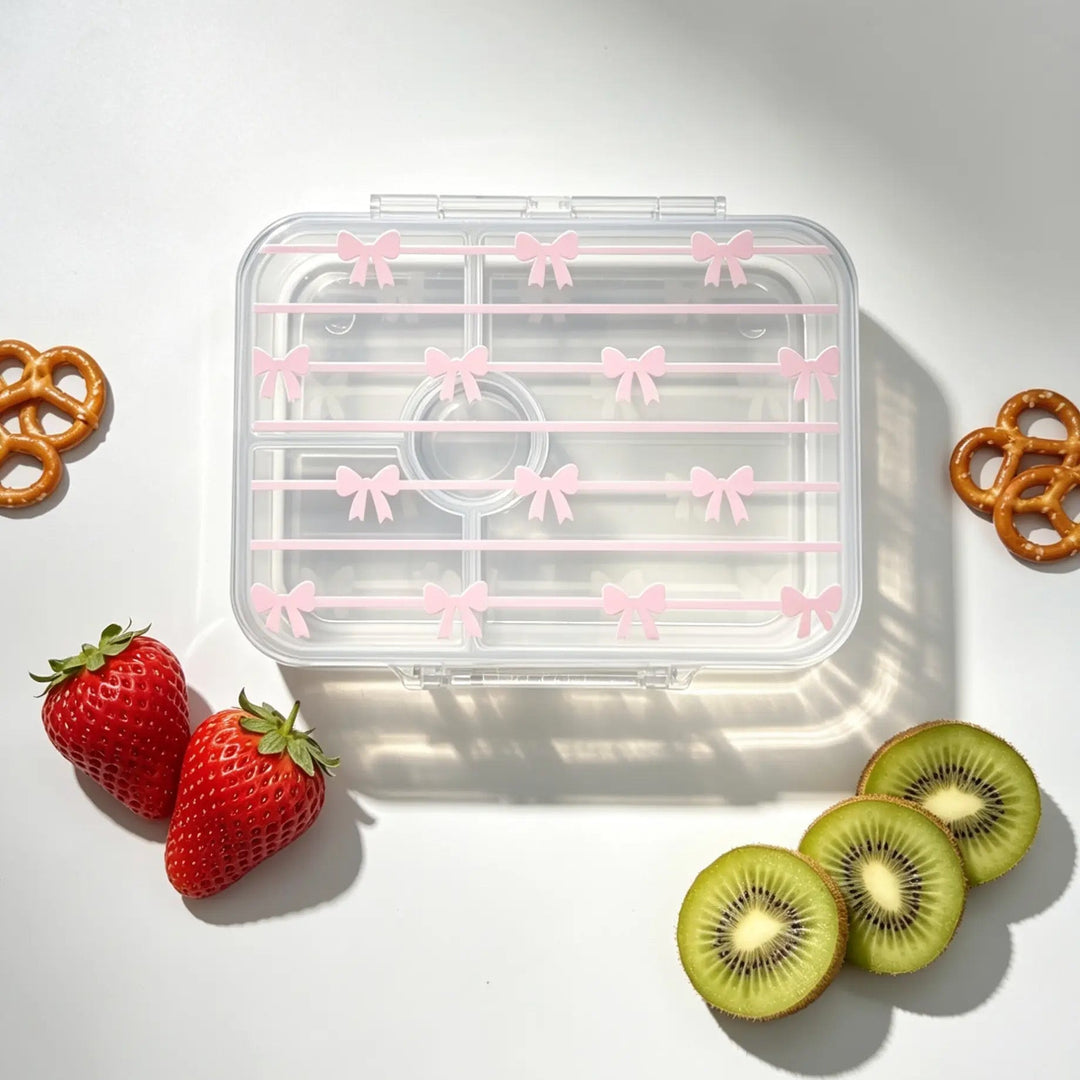 A clear plastic lunch box with a lid decorated with pink bow patterns, arranged on a white surface with decorative food props including strawberries, kiwi slices, and pretzels.