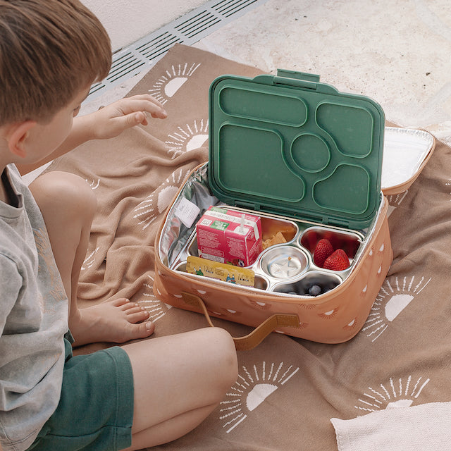 Child sitting on a patterned blanket with a bento box containing snacks.