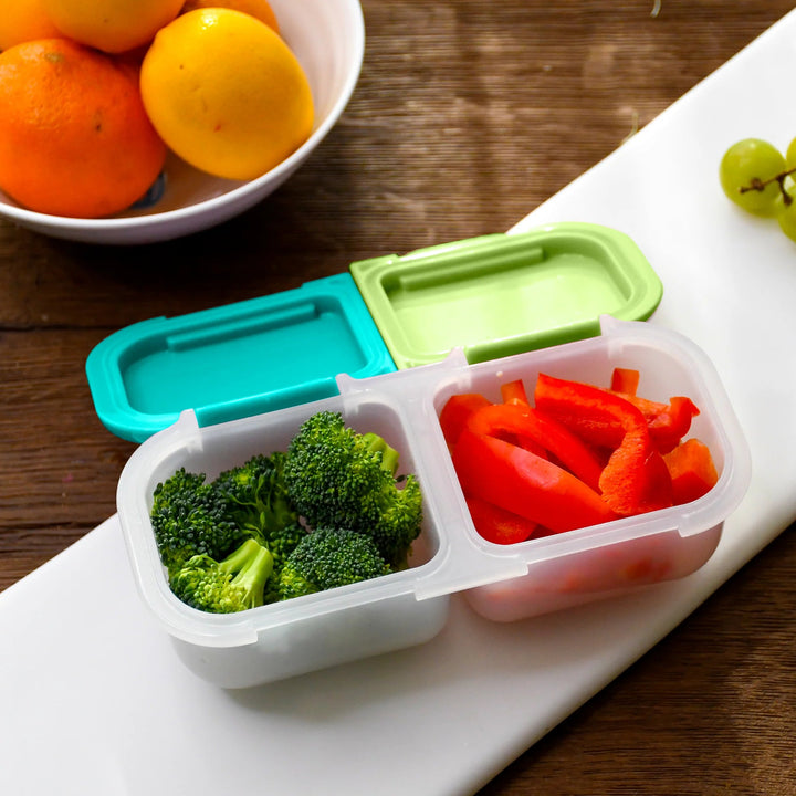 Meal prep container with compartments of broccoli and carrots on a wooden table with fruits in the background.